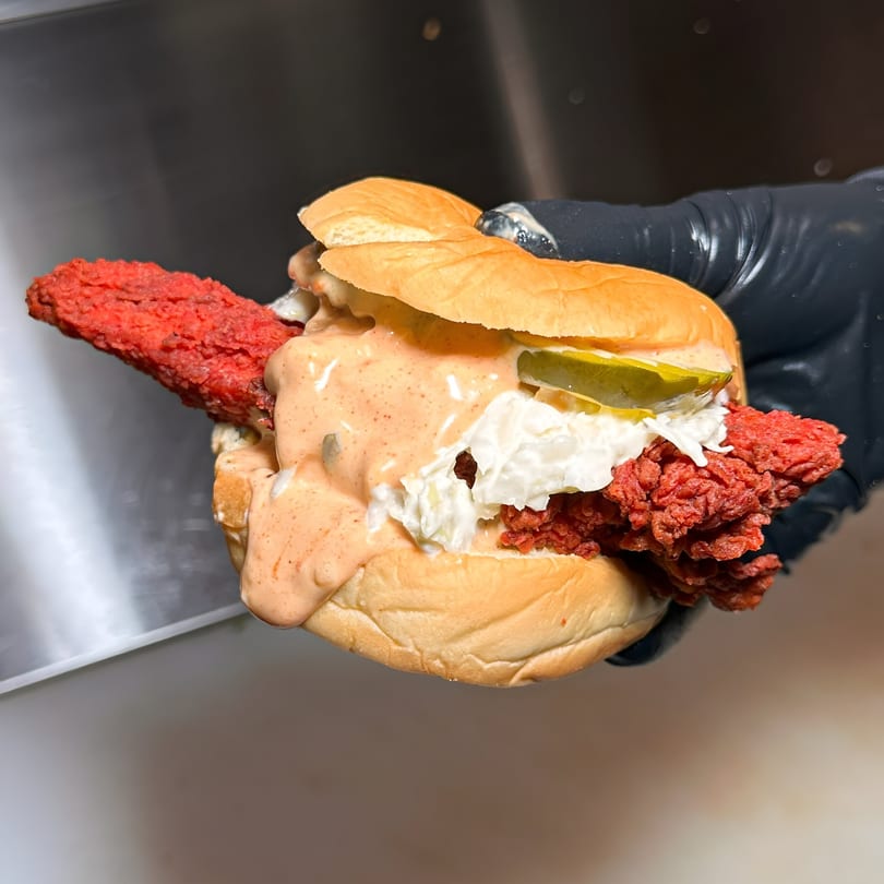 Hand in black glove holding a Chopped Cheese House sandwich with red meat, sauce, and pickles.