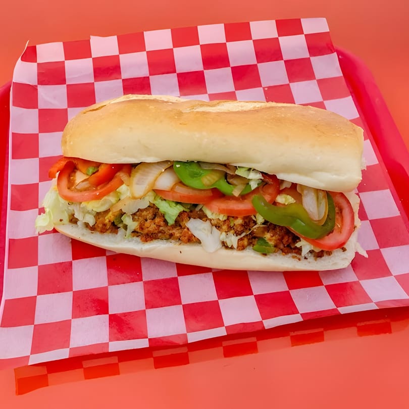 A Chopped Cheese House sandwich with meat, lettuce, tomato, onions, and peppers on a red checkered liner.